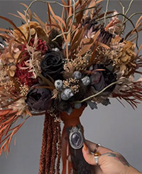 bouquet with black roses and orange natural toned flowers, leaves and branches, held by female hand on crystal wand with charm attached
