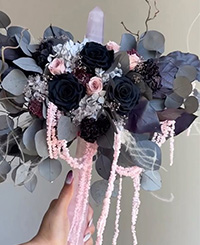 bouquet with black roses and light pink flowers surrounded by eucalyptus. Bouquet is draped with light pink floral pieces and held with female hand on crystal wand